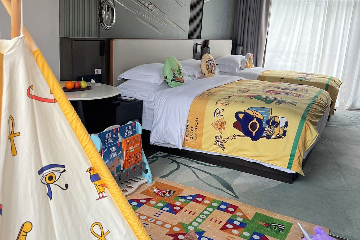 Una habitación de hotel con una cama, almohadas, una pequeña mesa con frutas, una alfombra colorida y una estructura parecida a una tienda con símbolos egipcios.