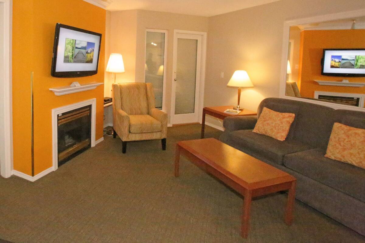 A hotel room with a bed, a sofa, a chair, a coffee table, a fireplace, and a television mounted on the wall.