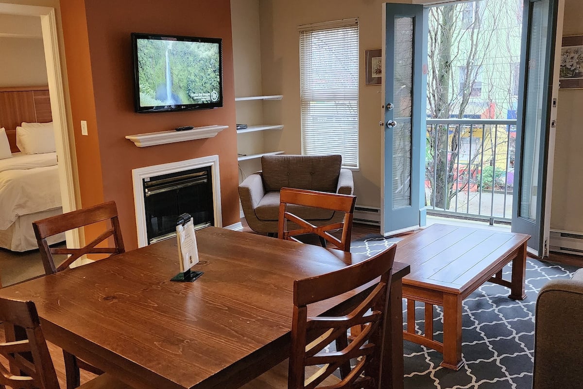 A hotel room with a bed, a TV mounted on the wall, a fireplace, a dining table with chairs, and a view of the outdoors through a door.