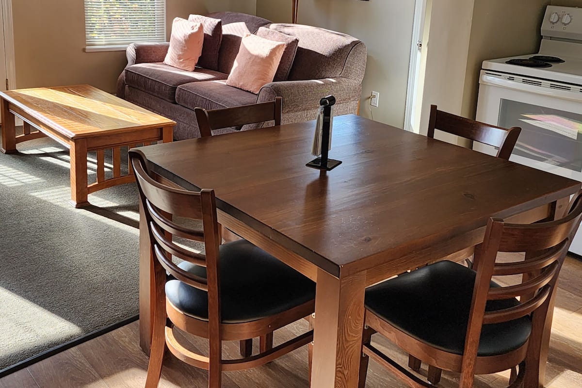 A cozy living room with a wooden dining table, cushioned chairs, a sofa with pillows, a lamp, and a stove.