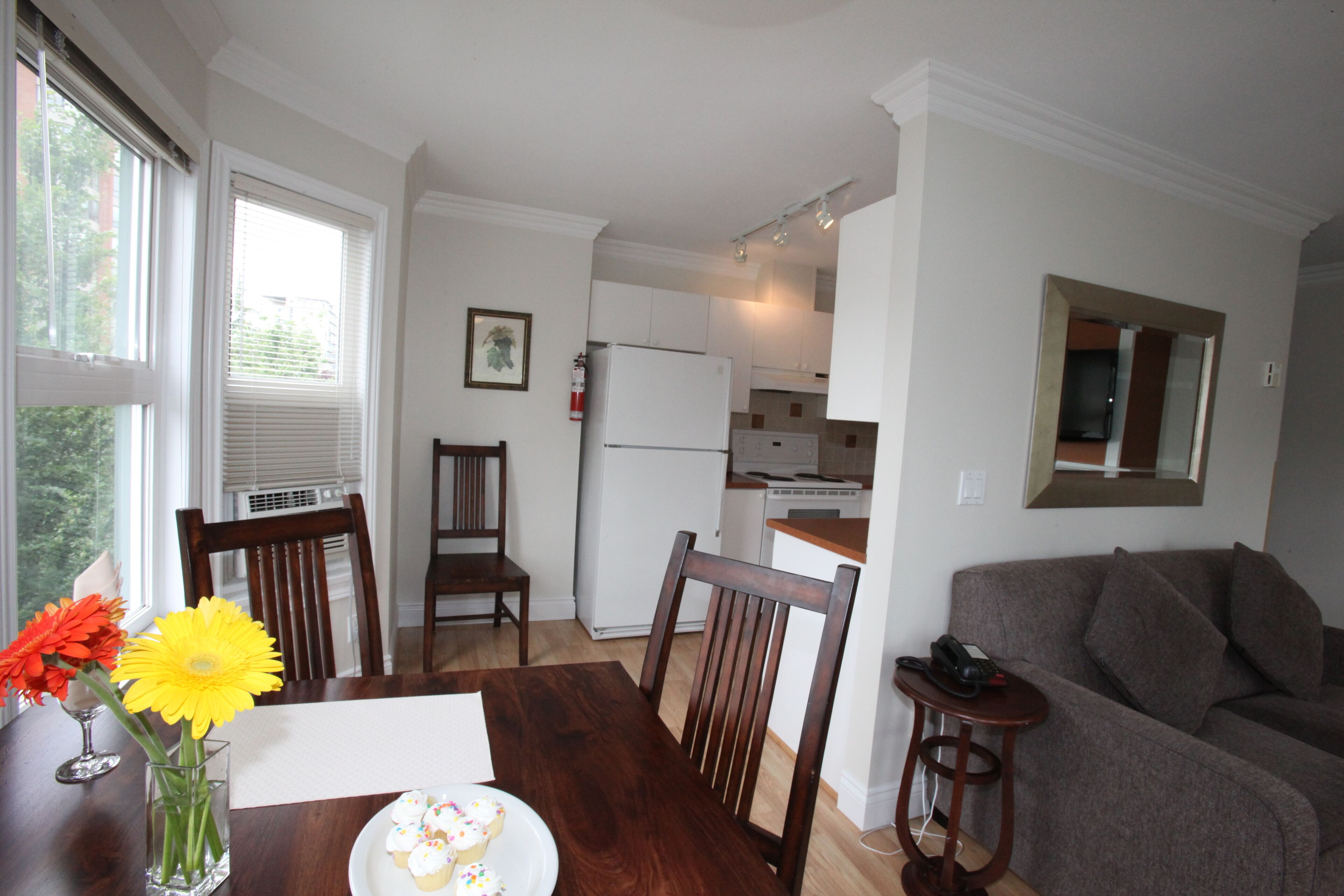 A dining area with a table, chairs, and a vase with flowers. There is a kitchen in the background with cabinets and appliances.