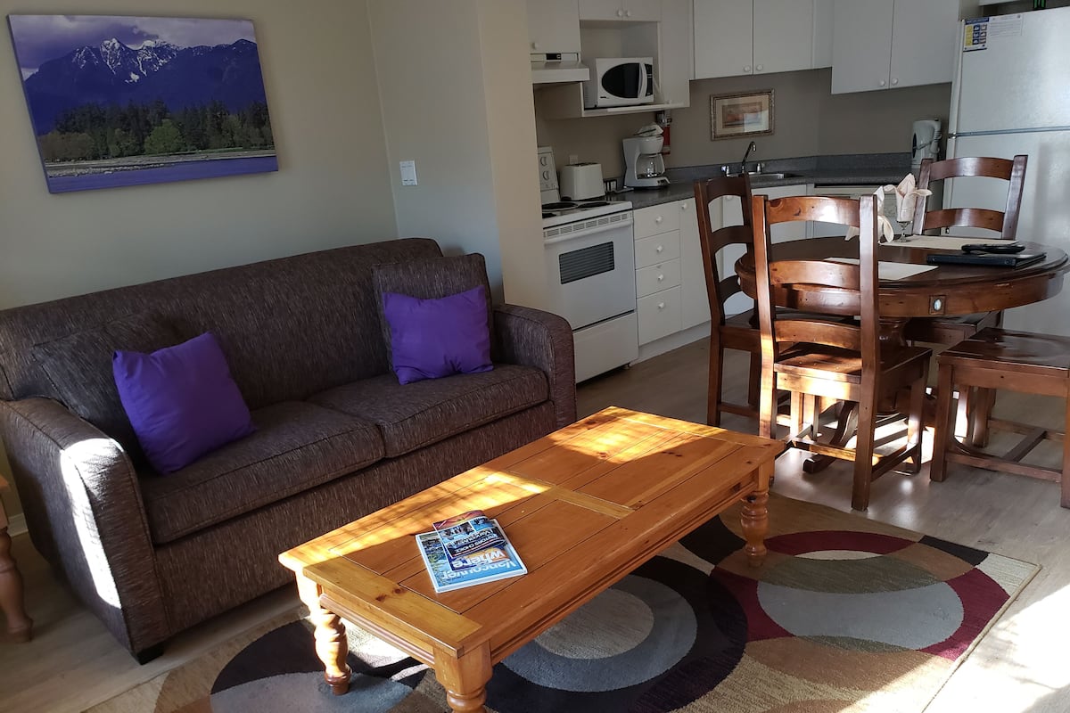 A living room with a brown sofa, purple cushions, a wooden coffee table, and a kitchen area with white cabinets and appliances.