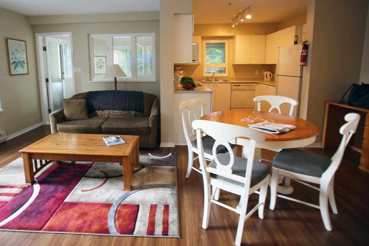 A modern living room with a sofa, coffee table, and dining table with chairs. There is a kitchen area with a refrigerator, stove, and sink.
