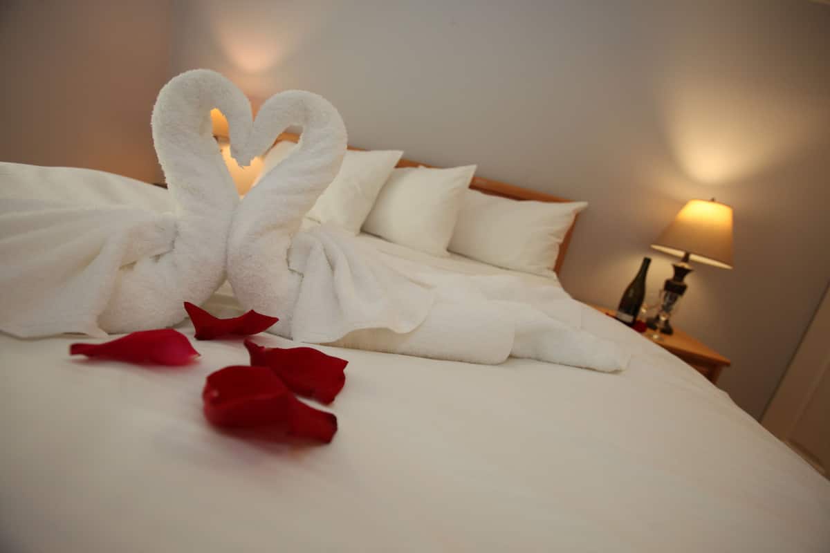 A neatly made bed with white linens and a towel arranged in the shape of a swan, along with red rose petals on the bed.