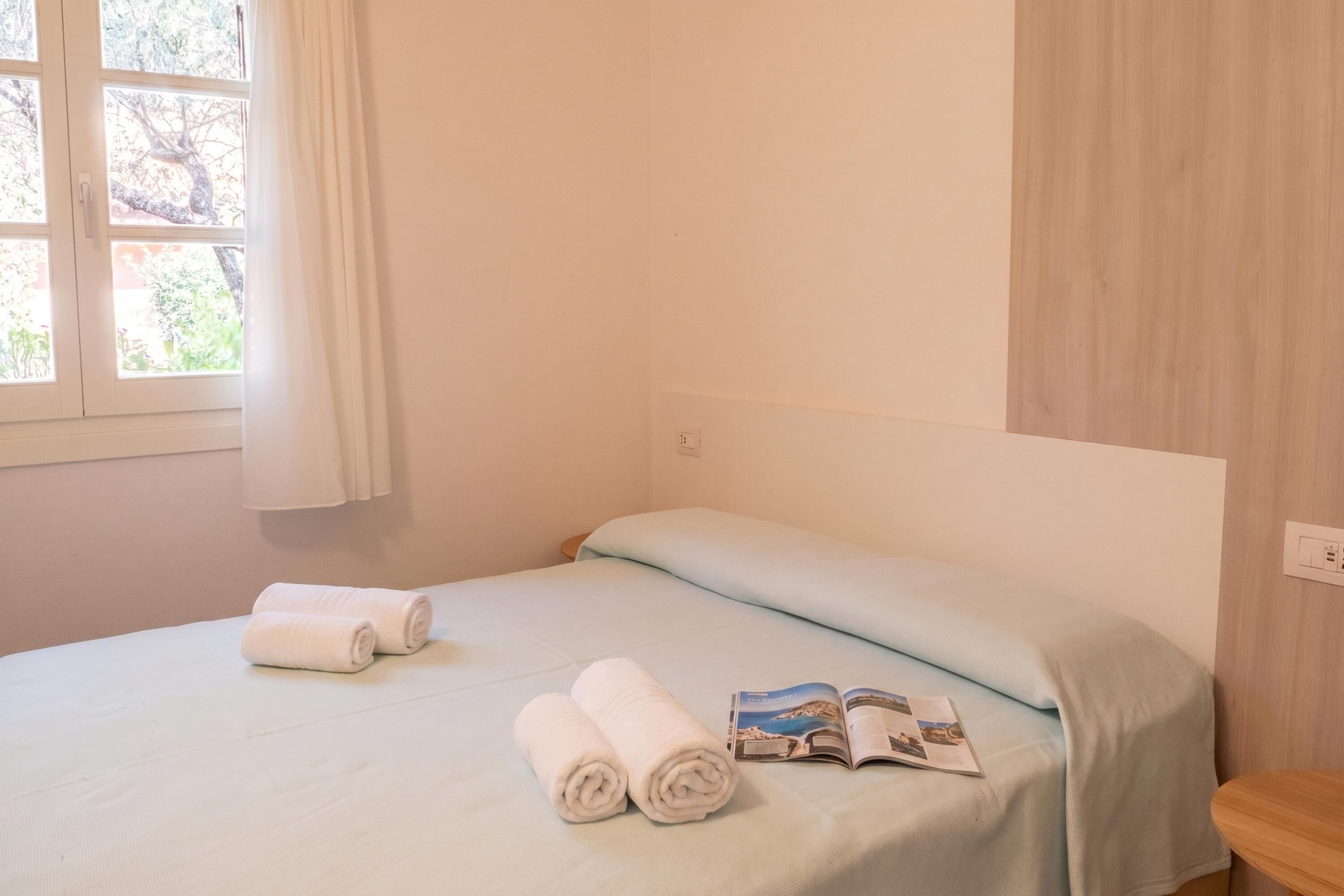 A single bed with a light blue bedspread, two rolled white towels, and a magazine on the bed.