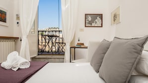 A hotel room with a bed, a small table with books, a window with a view of buildings, and a radiator. - Monti Deluxe - Terrace Spectacular View (Rome)
