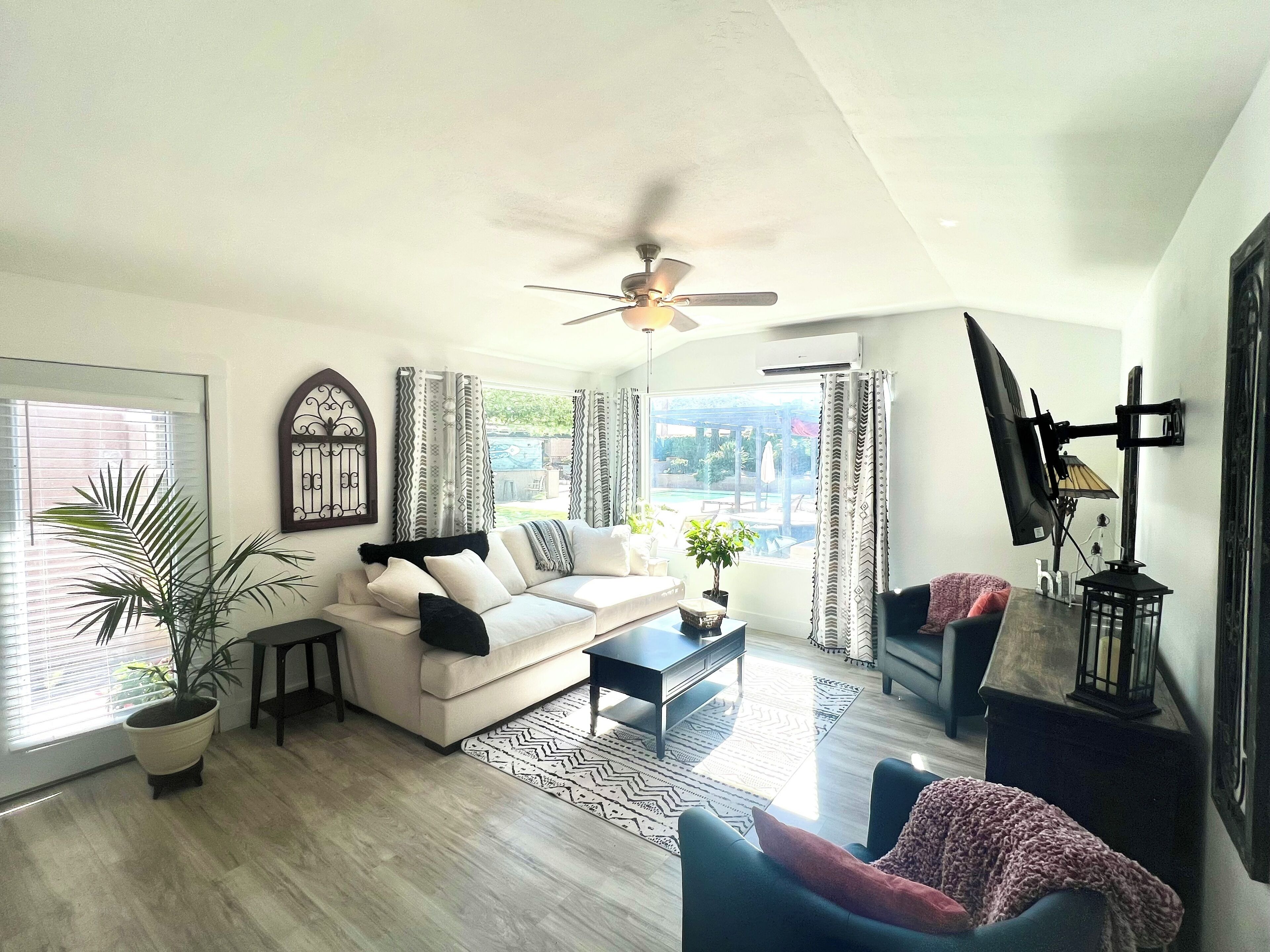 Ein modernes Wohnzimmer mit einer Couch, einem Couchtisch und einem Deckenventilator. Es gibt ein großes Fenster mit Vorhängen, einen Blumentopf und einen Blick ins Freie.
