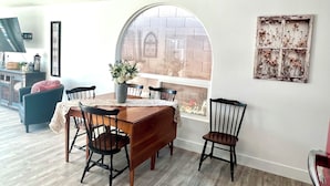 Un coin repas avec une table en bois, quatre chaises, un vase de fleurs, une grande fenêtre en arc de cercle et un élément décoratif fixé au mur.