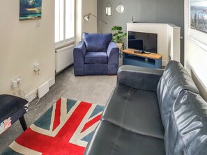 A living room with a blue sofa, a black leather sofa, a TV on a wooden stand, a radiator, and a window with a view of a snowy landscape. - 1 bedroom accommodation in Berwick-upon-Tweed (Berwick-upon-Tweed)
