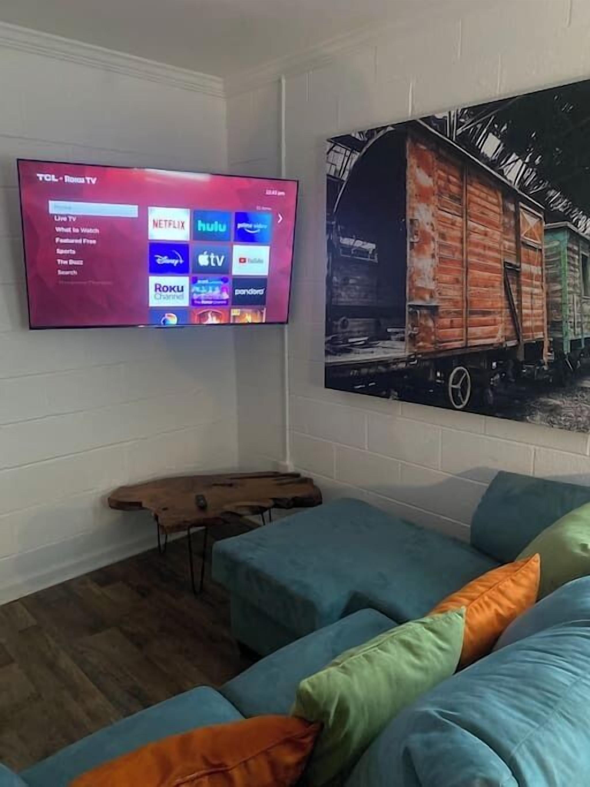 A living room with a flat-screen TV displaying streaming services, a blue sofa with colorful cushions, and a large painting of a train on the wall.