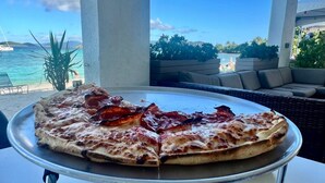Une pizza sur un plateau en métal, posée sur une table avec vue sur l’océan, et une terrasse en arrière-plan.