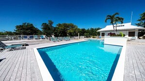 A rectangular swimming pool with a wooden deck surrounding it, a small building with a white roof, and palm trees in the background.