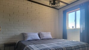 A bedroom with a bed, two pillows, a window with curtains, a ceiling fan, and a brick wall.