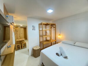 A hotel room with a bed, a wooden shelf unit, a small table with a chair, a mirror, and a wall-mounted light. - Flats no Beach Way - Praia dos Carneiros (Tamandaré)