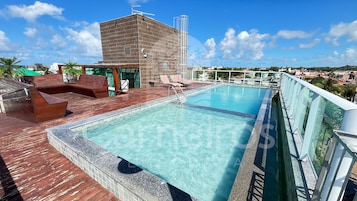 Una piscina en la azotea con agua cristalina, terraza de madera y sillones de descanso.