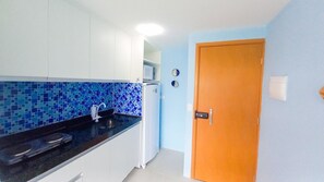 A kitchen with a black countertop, white cabinets, a blue mosaic backsplash, a white refrigerator, and a wooden door. - Flats no Beach Way - Praia dos Carneiros (Tamandaré)