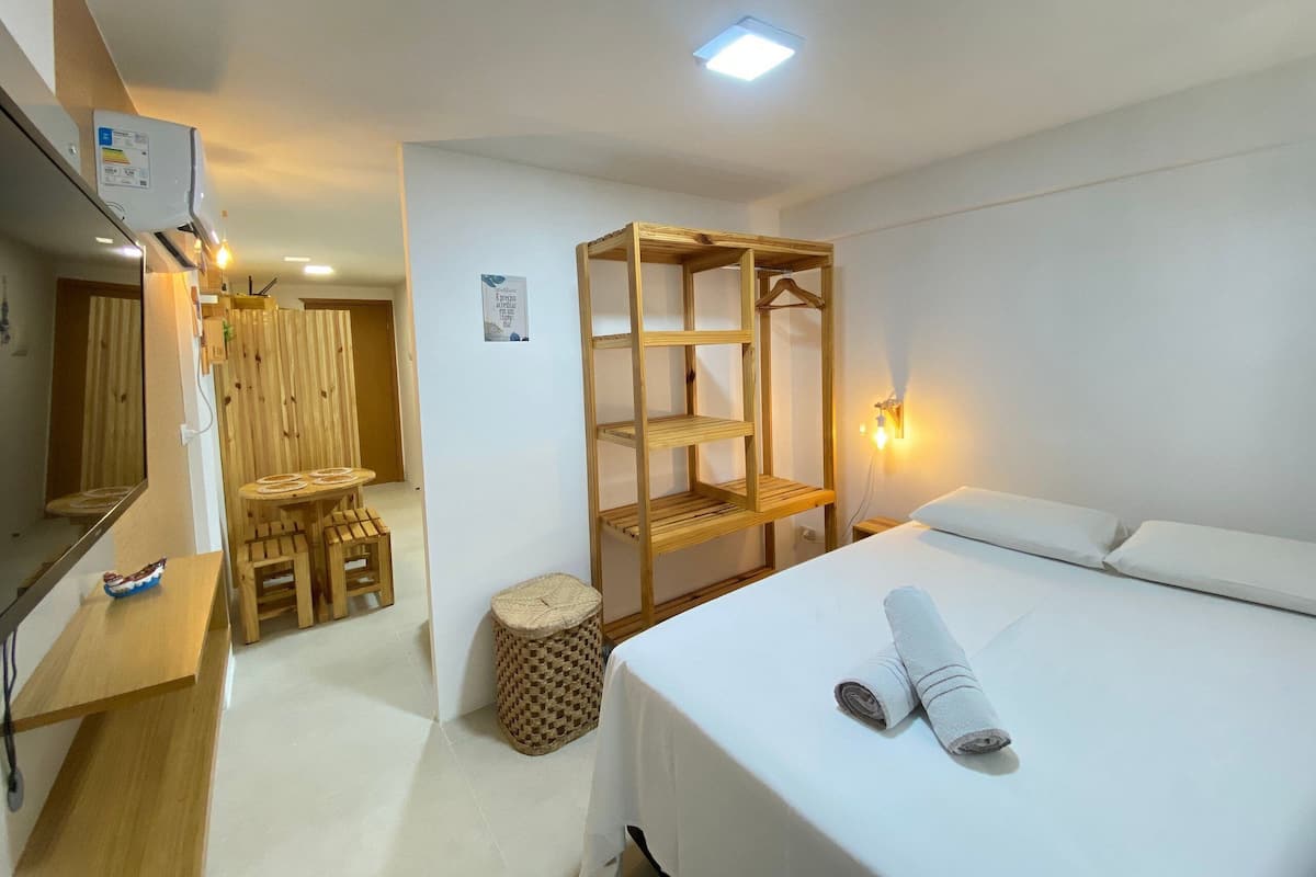 A hotel room with a bed, a wooden shelf unit, a small table with a chair, a mirror, and a wall-mounted light.