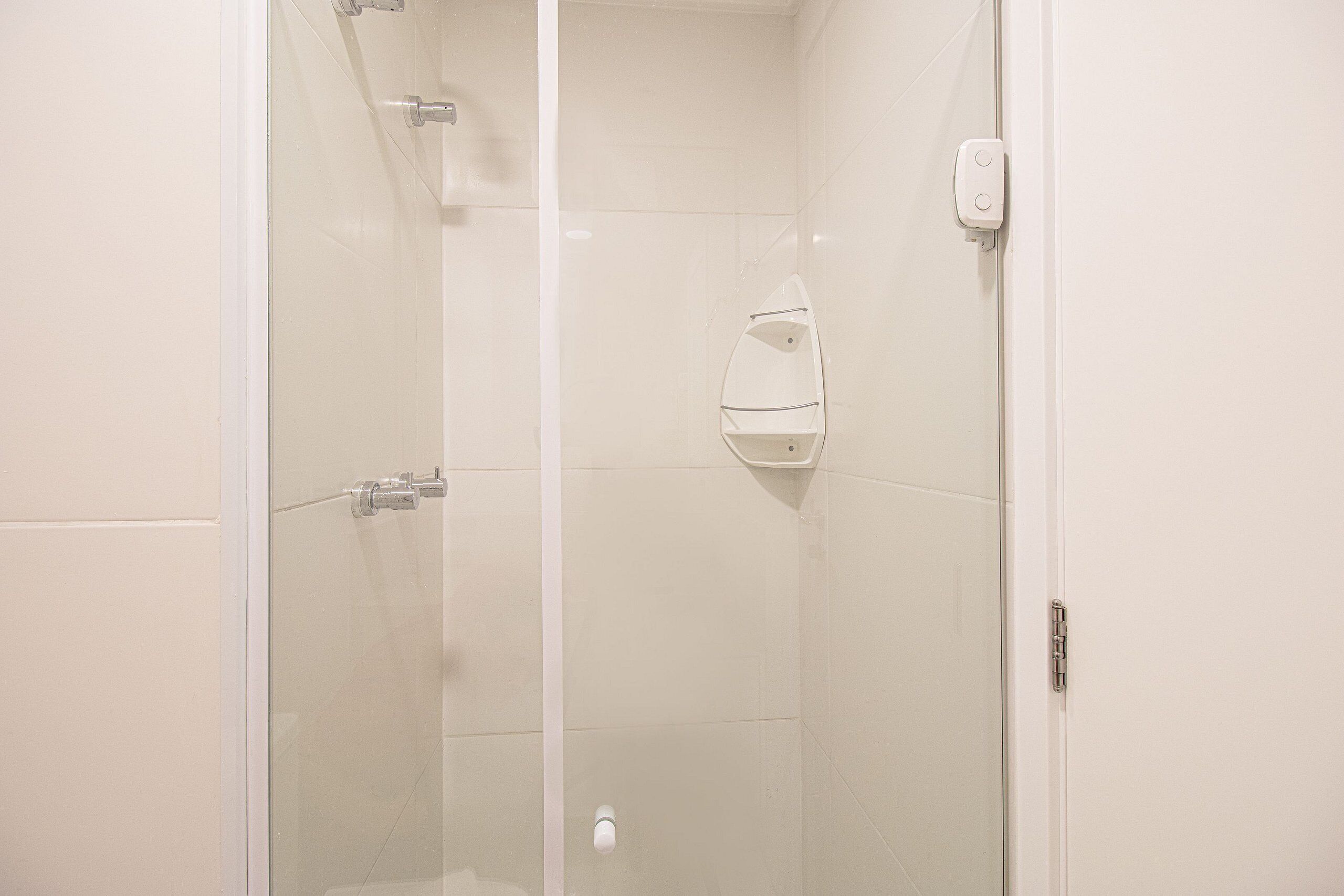 A glass-enclosed shower stall with a white tiled wall, a showerhead, and a soap dispenser.