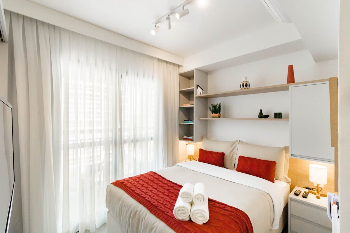 Ein Hotelzimmer mit Bett, Nachttisch, Lampe, einem Regal mit Dekoration und einem großen Fenster mit Vorhängen.