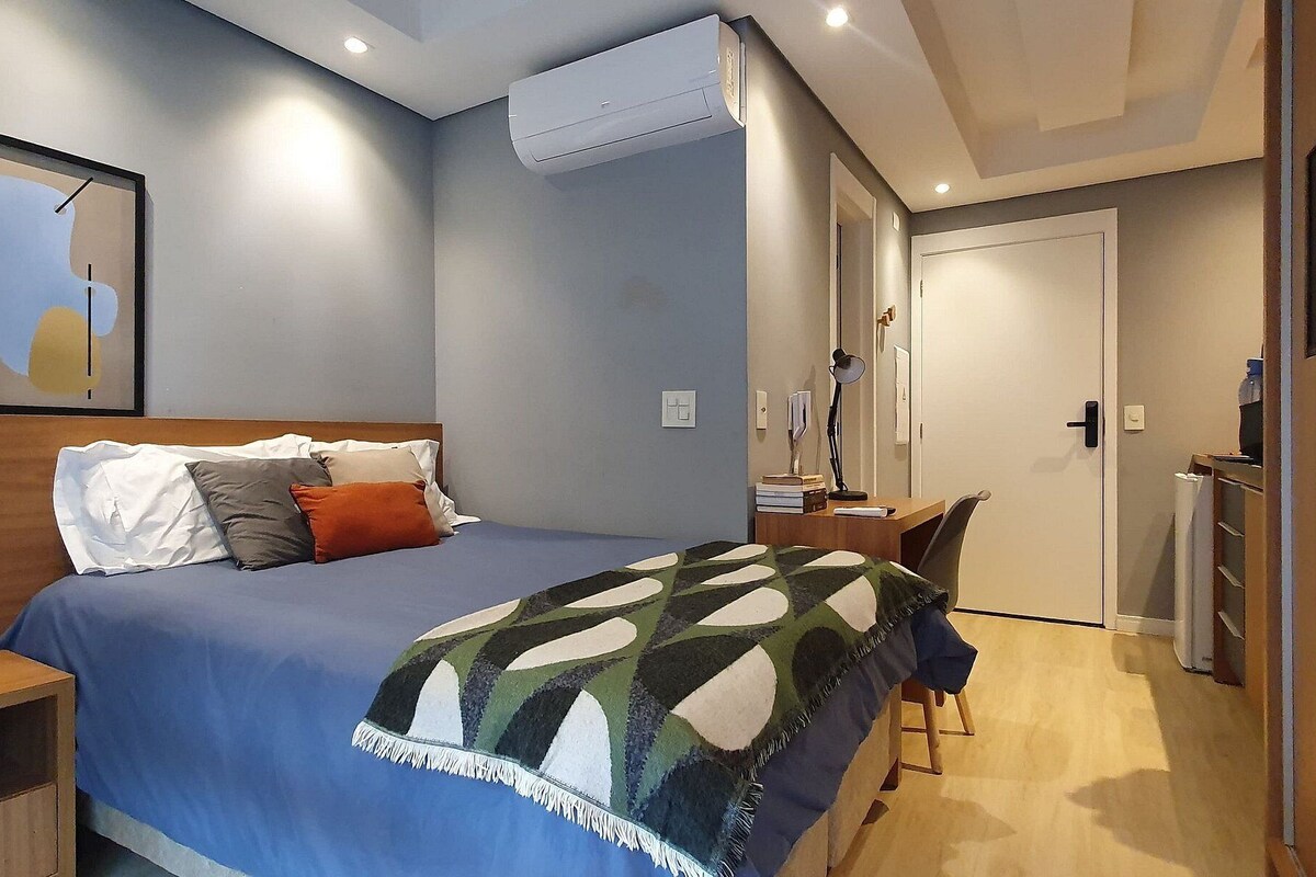 A bedroom with a bed, bedside table, desk, chair, and an air conditioning unit.