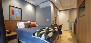 A bedroom with a bed, bedside table, desk, chair, and an air conditioning unit.