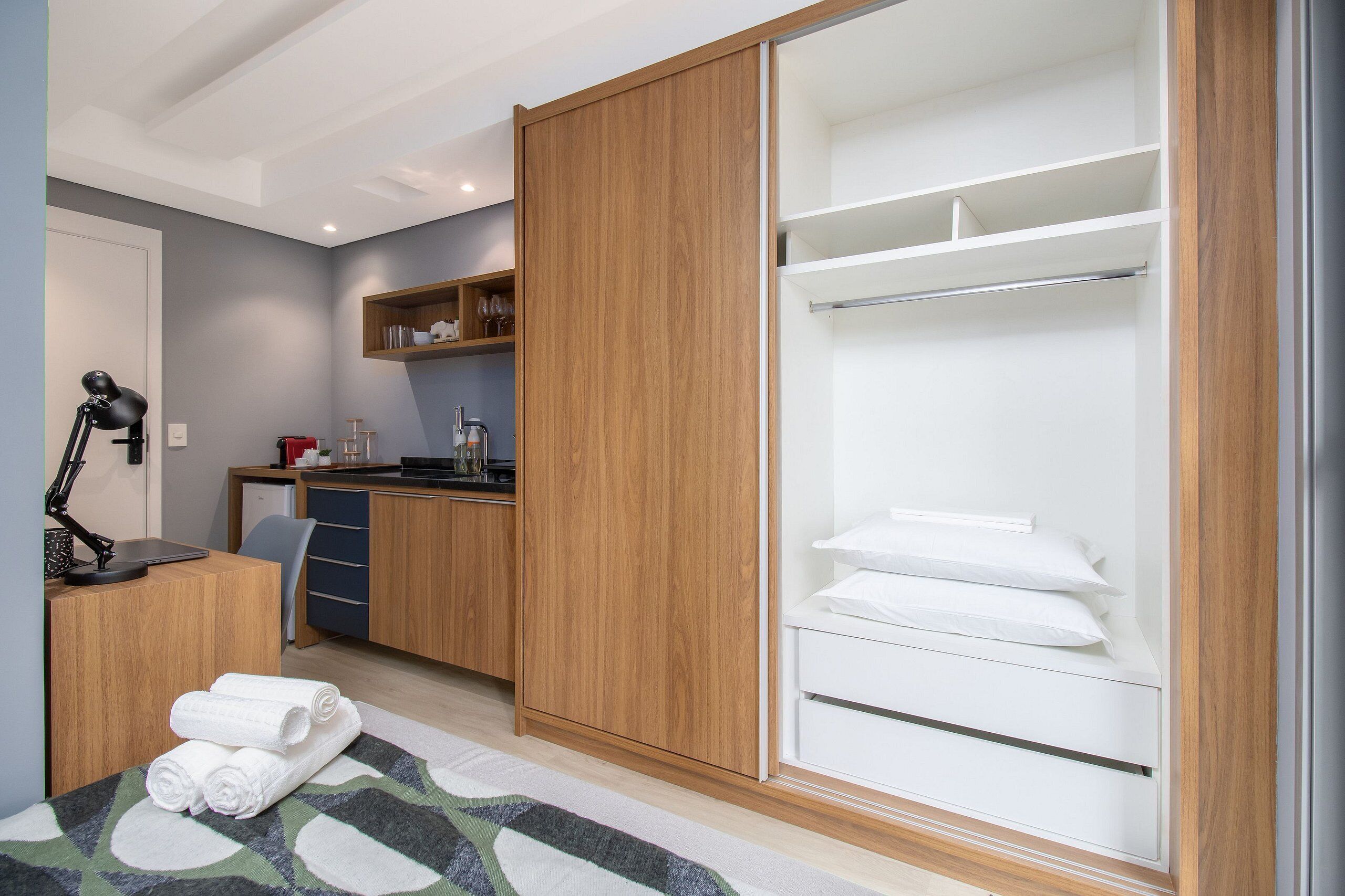 A hotel room with a wooden wardrobe, a desk with a lamp, a bed with rolled towels, and a kitchenette in the background.
