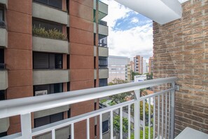 Un balcon doté d’une balustrade blanche, un mur en briques et une vue sur un immeuble moderne avec de nombreuses fenêtres et des balcons.