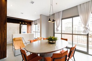 A modern conference room with a round table, orange chairs, a potted plant, and large windows with curtains.