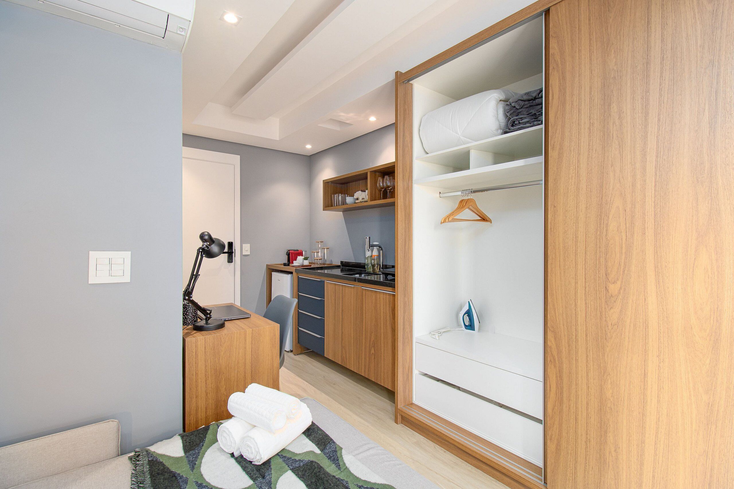 A compact room with a bed, a desk with a lamp, a kitchenette, and a wardrobe with hanging space.