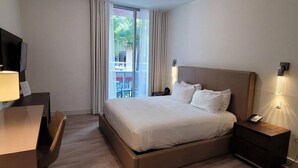 A hotel room with a large bed, a desk, a chair, a TV, and a balcony with curtains.