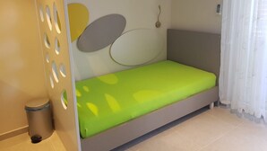 Ein kleines Zimmer mit einem Einzelbett, einer grünen Matratze, einer Wandleuchte und einem Fenstervorhang.