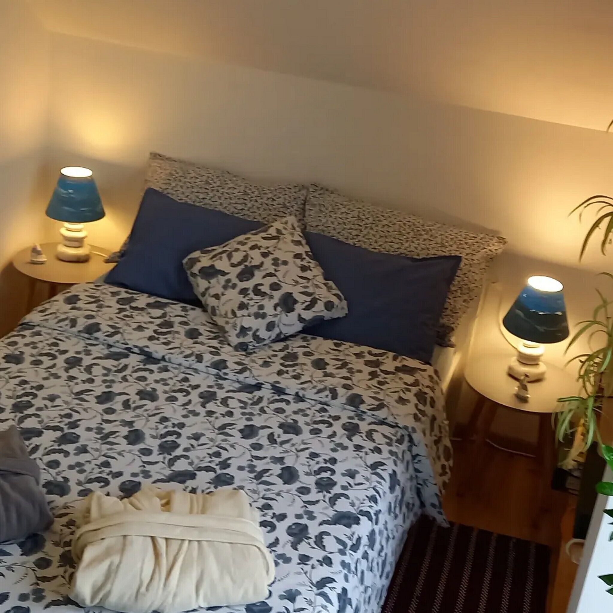 Siististi pedattu sänky, jossa on kukkakuviollinen peitto ja tyynyjä, kaksi yöpöytävalaisinta ja yöpöydällä oleva kasvi.