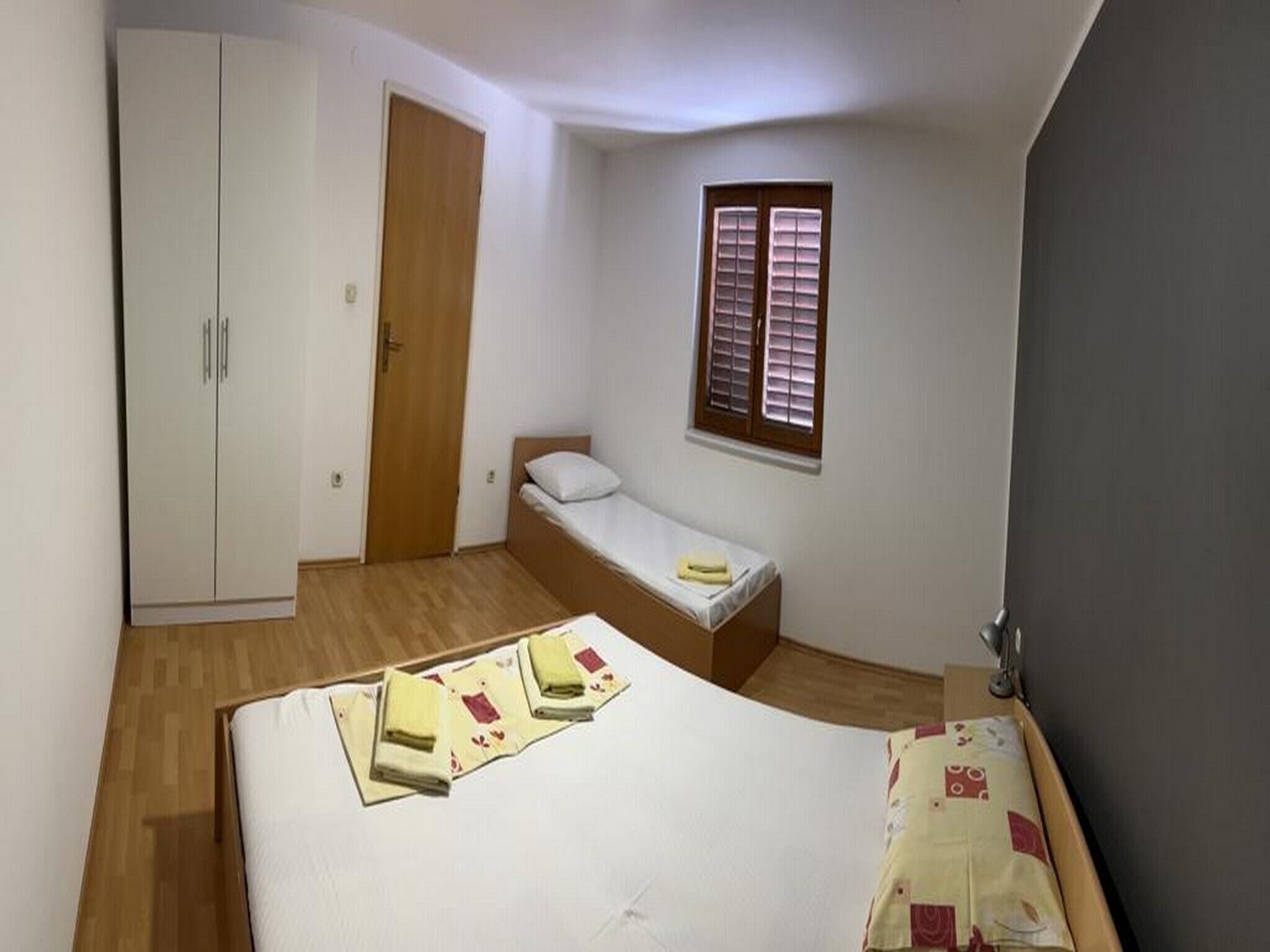 A small bedroom with a single bed, a wardrobe, a window with blinds, and a door.