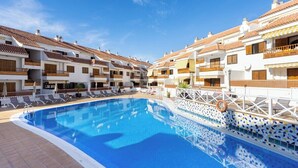 A swimming pool surrounded by white buildings with balconies, a tiled deck, and a clear blue sky.