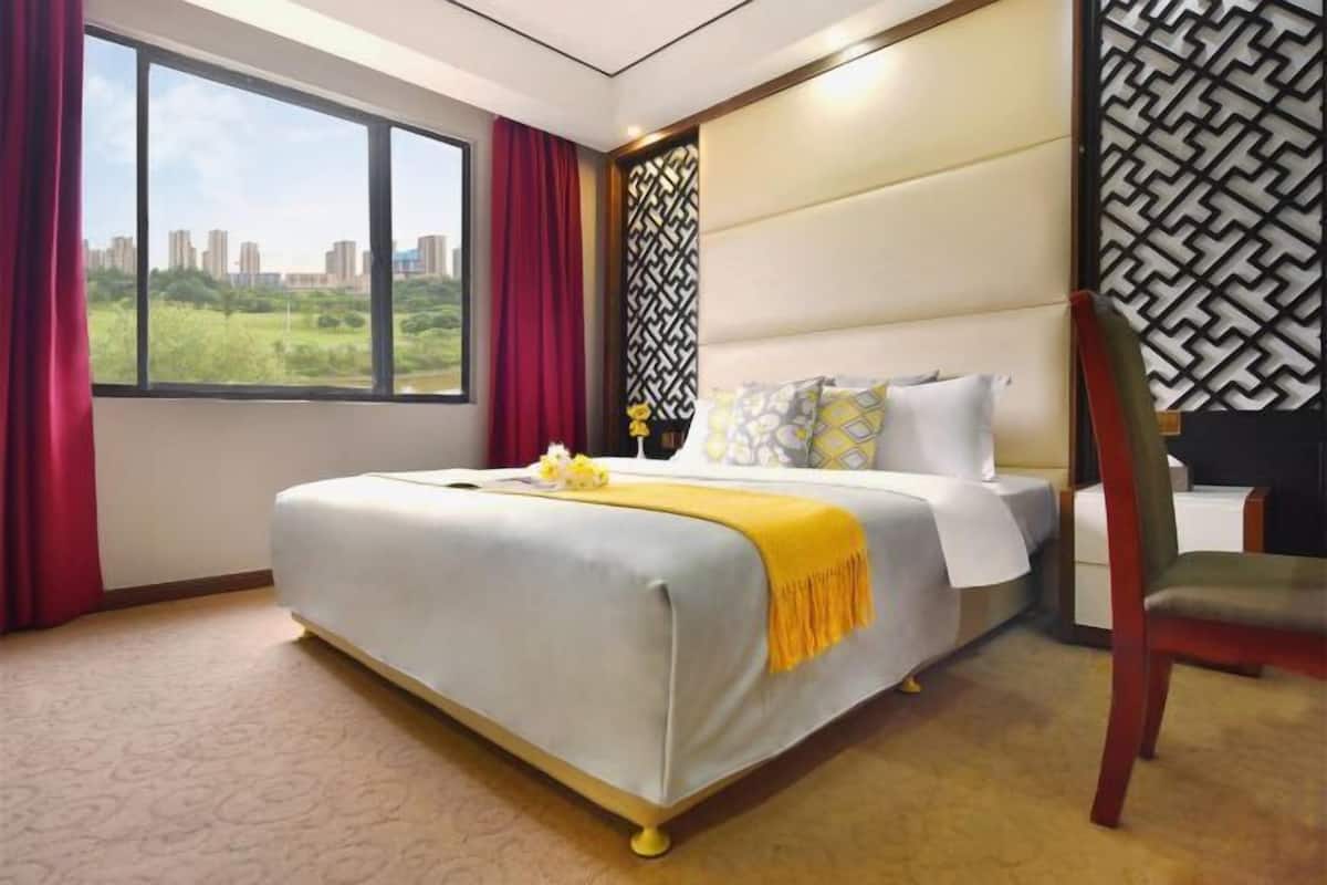 A hotel room with a large bed, a view of greenery and buildings, and a patterned wall.