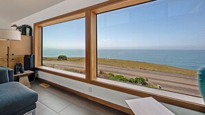 A room with large windows overlooking a coastal road and ocean, featuring a desk, chairs, and a lamp. - Ocean Atrium - Amazing Frontline Views (Bodega Bay)
