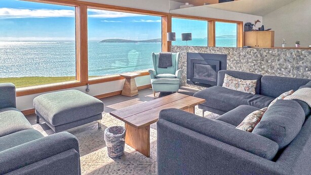 A modern living room with a large window overlooking the sea, a fireplace, and comfortable seating. - Ocean Atrium - Amazing Frontline Views (Bodega Bay)