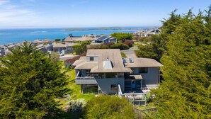 A coastal residential area with houses, some with solar panels, surrounded by greenery and overlooking the sea.