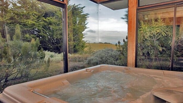 A hot tub with a control panel, surrounded by glass walls offering a view of a garden and trees.