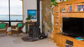 A cozy living room with a stone fireplace, a large flat-screen TV, wicker furniture, and a view of the sea through large windows. - Boccabella - Quite south side location, expansive views. (Bodega Bay)
