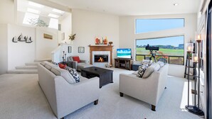 A spacious living room with a fireplace, comfortable seating, and a large window offering a view of the outdoors. - Dabbling Duck - Light, open, warm home on the 14th fairway, great views (Bodega Bay)