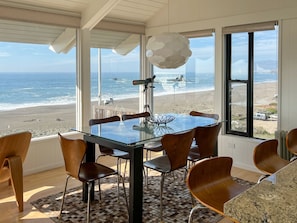 A dining area with a glass table and wooden chairs, overlooking a beach with waves and a rocky outcrop. - Louw Tide - Modern home, ocean front, spanning views. (Bodega Bay)