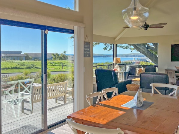 A spacious room with a wooden table and chairs, large windows offering a view of the sea and greenery, and a ceiling fan. - Paradise on the Bay - Nice ocean views, 1 level (Bodega Bay)