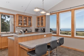 A modern kitchen with wooden cabinets, a central island with seating, and large windows offering ocean views.