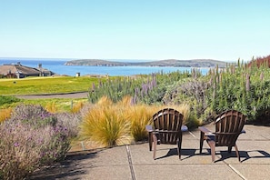Zwei hölzerne Stühle auf einer Terrasse mit Blick auf eine Küstenlandschaft, Meer und Hügel.