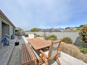 A wooden deck with a table and chairs, surrounded by a garden with purple flowers and a wooden fence.