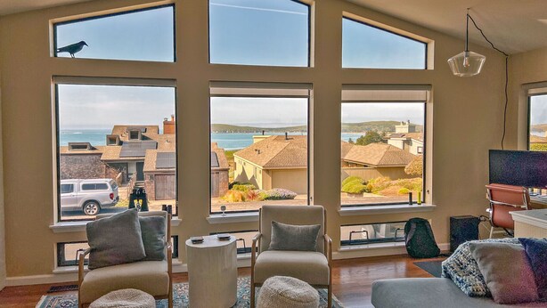A spacious living room with large windows, a cozy seating area, and a view of the sea and buildings.
