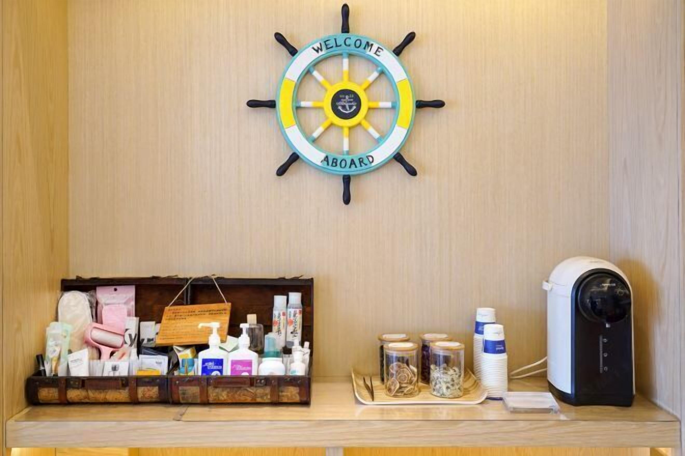 A wooden wall with a ship's wheel sign saying "WELCOME ABOARD" and a wooden shelf with toiletries and a coffee machine.