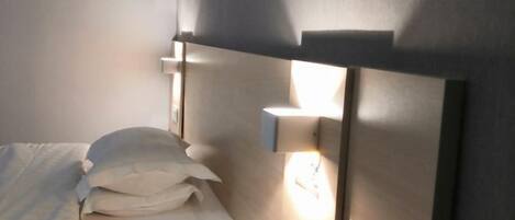 Un lit avec une literie et des oreillers blancs, une table de chevet et un luminaire fixé au mur.