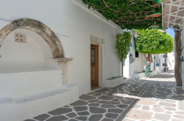 A narrow stone-paved street with white buildings, a wooden door, and a tree with hanging decorations. - 2-Storey Cycladic House In Old Town Sleeps 20 (Paros)